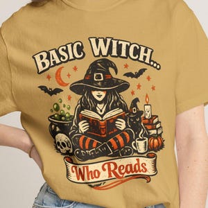May include: A mustard yellow t-shirt featuring a graphic print of a witch reading a book. The text reads "BASIC WITCH... Who Reads". The illustration includes a cauldron, a skull, a black cat, a pumpkin, a candle, bats, and a crescent moon.