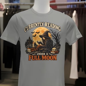 May include: A grey short-sleeve t-shirt featuring a graphic print of a witch reading a book under a large yellow full moon. The text reads CURRENTLY READING UNDER A FULL MOON. The design includes a black cat, a skull, a candle, and bats.