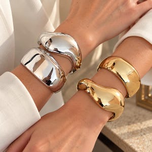 Op de afbeelding: Twee zilverkleurige metalen armbanden met een gladde, onregelmatige, organische vorm om de ene pols, en twee goudkleurige metalen armbanden met een vergelijkbare gladde, onregelmatige, organische vorm om de andere pols, alle met een hoogglans afwerking.