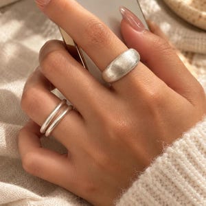 Op de afbeelding: Een brede, geborstelde zilverkleurige ring aan de middelvinger en een set van twee dunne, gladde zilverkleurige ringen aan de ringvinger. De ringen hebben een matte, metalen afwerking en een eenvoudig, ongekunsteld ontwerp.