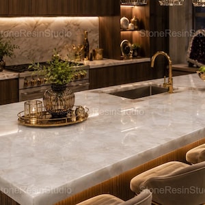 Puede incluir: Una isla de cocina con encimera de cuarzo blanco pulido con vetas grises sutiles, un grifo dorado y un fregadero. Sobre la isla hay una bandeja dorada con un portavelas de cristal y un jarrón de cristal oscuro con follaje verde.