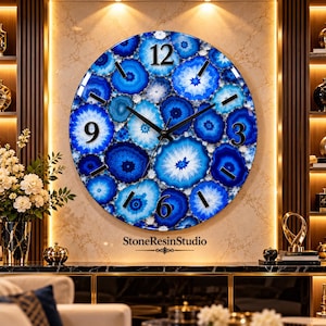 May include: A round wall clock featuring a vibrant blue agate slice pattern across the face. The clock has black hands and black numerals at the 12, 3, 6, and 9 positions. The text StoneResinStudio is printed in black below the clock face.