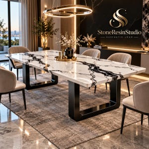 Op de afbeelding: Een rechthoekige eettafel met een wit marmeren blad met opvallende zwarte aders, ondersteund door twee dikke, zwarte U-vormige metalen poten. De tafel is omringd door vier beige gestoffeerde stoelen met zwarte poten.