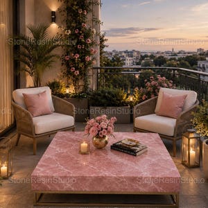 May include: A square coffee table with a pink marble top and a gold metal base, positioned between two beige woven armchairs with pink cushions. A gold vase with pink flowers and a stack of books sit on the table, flanked by two lanterns.