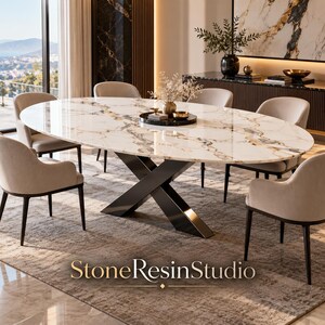 Puede incluir: Una mesa de comedor ovalada con tablero de mármol blanco con vetas grises y doradas, apoyada sobre una base de metal negro en forma de X. Seis sillas tapizadas en beige con patas oscuras rodean la mesa. El texto StoneResinStudio aparece abajo.