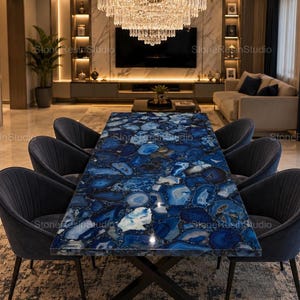 Puede incluir: Una mesa de comedor rectangular con una superficie de ágata azul pulida que presenta un mosaico de rodajas de piedra en tonos azules y blancos, apoyada sobre una base de metal oscuro en forma de X, rodeada de seis sillas tapizadas en azul.