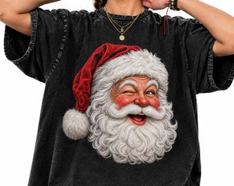 Può includere: Una t-shirt nera con effetto lavaggio acido caratterizzata da una grande stampa grafica del volto di Babbo Natale che fa l'occhiolino. Il disegno mostra una folta barba bianca, guance rosate e un cappello rosso con un pon-pon bianco.
