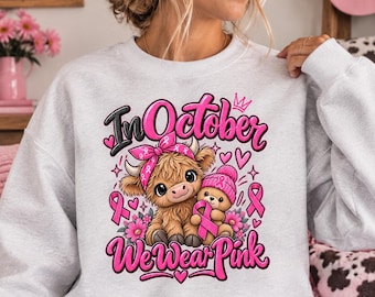 Può includere: Una felpa girocollo grigio chiaro con una stampa grafica di una soffice mucca Highland con una bandana rosa e un orsacchiotto con un berretto rosa. Il testo recita In October We Wear Pink, circondato da nastri rosa, cuori e fiori.