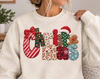 Può includere: Una felpa girocollo color crema con la scritta Christmas in grandi lettere a motivi vari. Il design include strisce rosse e bianche tipo bastoncino di zucchero, tartan verde, stampa leopardata, pois rossi, renne e un omino di pan di zenzero.