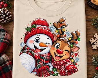 May include: A beige t-shirt featuring a graphic illustration of a winking snowman and a winking reindeer. The snowman wears a red knit hat and scarf, while the reindeer has antlers wrapped in colorful Christmas lights and a red scarf.