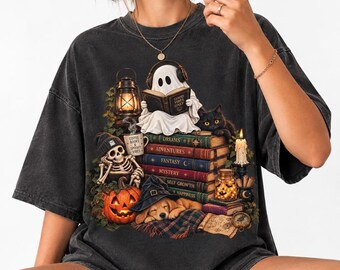 Può includere: Una t-shirt grigio antracite con una stampa grafica di un fantasma con le cuffie che legge un libro intitolato Good Vibes Only, uno scheletro, un gatto nero, una pila di libri, una zucca e un cucciolo con un cappello da strega.