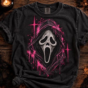 May include: A black short-sleeve t-shirt featuring a central graphic of a white ghost-like mask with a wide open mouth, surrounded by a hooded cloak. The design is accented with bright pink paint splatter effects and four-pointed star shapes.