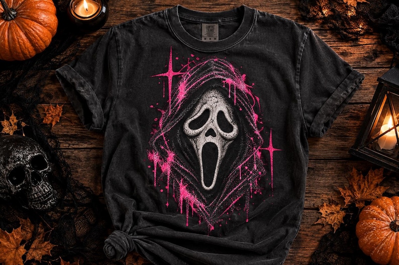 May include: A black short-sleeve t-shirt featuring a central graphic of a white ghost-like mask with a wide open mouth, surrounded by a hooded cloak. The design is accented with bright pink paint splatter effects and four-pointed star shapes.