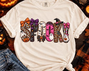 Può includere: Una t-shirt color crema con la scritta SPOOKY in grandi lettere decorative. Ogni lettera è riempita con motivi a tema Halloween, tra cui stampa leopardata, fantasmi, zucche, pipistrelli, ragnatele e fiocchi, sormontata da un cappello da strega.