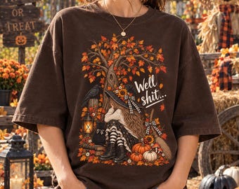 Può includere: Una t-shirt a maniche corte marrone scuro con una stampa grafica di una persona con un cappello a tesa larga seduta sotto un albero autunnale con foglie arancioni. Il design include un corvo, una lanterna, zucche e il testo "Well shit..." in corsivo bianco.