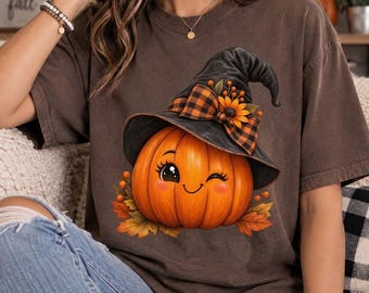 Può includere: Una t-shirt a maniche corte marrone scuro con la grafica di una zucca arancione che fa l'occhiolino, indossando un cappello da strega nero. Il cappello è decorato con un fiocco a quadri arancione e nero, un girasole e piccole bacche.
