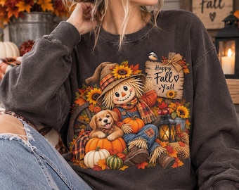 Può includere: Una felpa girocollo grigio antracite con la grafica di uno spaventapasseri con sciarpa e cappello a quadri, seduto con un cucciolo dorato tra zucche, girasoli e una lanterna. Il testo Happy Fall Y'all è scritto su un cartello di legno.