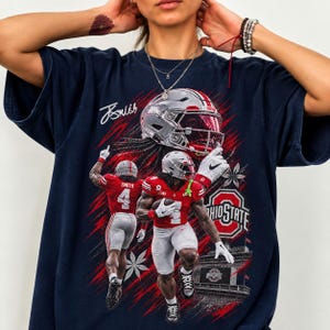 May include: A navy blue t-shirt featuring a graphic print of Ohio State football players in red uniforms with white helmets. The design includes a large helmet, the text "Smith" with number 4, the Ohio State logo, and a stylized signature.