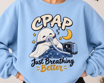 Può includere: Una felpa girocollo azzurro chiaro con la grafica di un fantasma bianco che indossa una maschera CPAP e un tubo per la respirazione, coperto da una coperta blu con stelle. Il testo recita CPAP Just Breathing Better con una luna gialla.