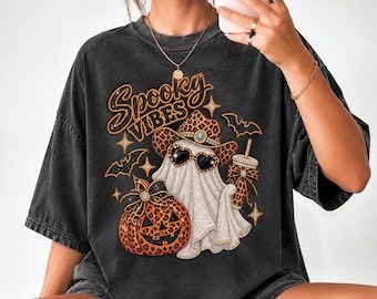 Può includere: Una t-shirt oversize grigio antracite con una stampa grafica di un fantasma che indossa un cappello da cowboy e occhiali da sole a forma di cuore con stampa leopardata. Il fantasma tiene un bicchiere leopardato accanto a una zucca. Testo: Spooky Vibes.