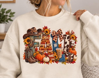 Può includere: Una felpa girocollo color crema con una stampa grafica della parola FALL. Le lettere sono decorate con un cappello da cowboy, girasoli, un fiocco a quadri, un bicchiere di caffè con scritto Fall Vibes, zucche e foglie autunnali.