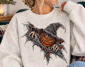 Può includere: Una felpa girocollo color beige chiaro con una stampa grafica raffigurante una zucca di Halloween arancione brillante che indossa un cappello da strega nero logoro, con mani scheletriche che emergono da un effetto di tessuto strappato sul davanti.
