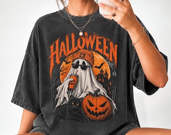 Può includere: Una t-shirt oversize grigio antracite con una stampa grafica che riporta la scritta HALLOWEEN in arancione. Il disegno raffigura un fantasma con berretto e occhiali da sole che tiene una bevanda, un gatto nero, una zucca e una casa stregata.