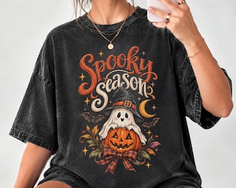 Può includere: Una t-shirt nera lavata al minerale con una stampa grafica che riporta il testo "Spooky Season" in corsivo arancione. Il design include un fantasma con cappello da strega su una zucca, circondato da pipistrelli, stelle e un fiocco a quadri.