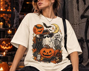 Può includere: Una t-shirt a maniche corte color bianco sporco con una stampa grafica colorata a tema Halloween. Il design include un fantasma con cappello e occhiali da sole, un gatto nero, uno scheletro, una zucca, pipistrelli, una luna e una casa.