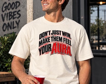 Può includere: Una t-shirt a maniche corte color crema con testo nero e rosso sul petto che recita "DON'T JUST WIN, MAKE THEM FEEL YOUR AURA." La maglietta presenta un girocollo e una vestibilità standard, realizzata in un tessuto testurizzato.