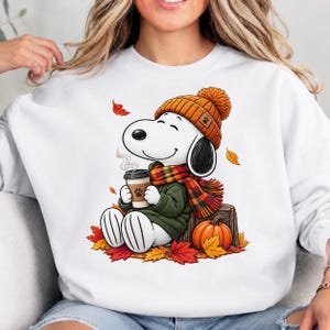 Puede incluir: Una sudadera blanca de cuello redondo con un gráfico de un perro de dibujos animados blanco que lleva un gorro de punto naranja, un suéter verde y una bufanda a cuadros. El perro sostiene una taza de café sentado en un tronco junto a una calabaza.