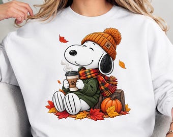 May include: A white crewneck sweatshirt featuring a graphic of a white cartoon dog wearing an orange knit beanie, a green sweater, and a plaid scarf. The dog holds a coffee cup while sitting on a tree stump next to an orange pumpkin and autumn leaves.