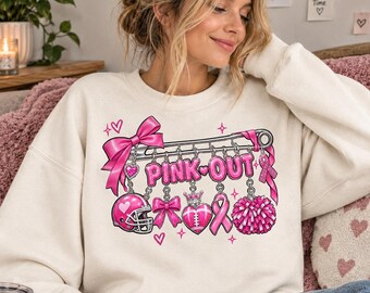 Può includere: Una felpa girocollo color crema con la grafica di una grande spilla da balia argentata e la scritta PINK OUT. Dalla spilla pendono ciondoli rosa tra cui un casco da football, fiocchi, un cuore, un nastro e un pon pon da cheerleader.