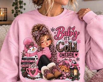 May include: A light pink crewneck sweatshirt featuring a graphic of a cartoon character with a leopard print bow, a snowman, a mug of hot cocoa, a puppy, and a lantern. The text reads Baby It's Cold Outside with stacked books labelled Hot Cocoa, Cozy Vibes, Warm Hearts, and Cold Days.