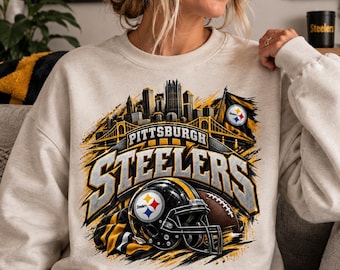 Könnte beinhalten: Ein beigefarbenes Sweatshirt mit Rundhalsausschnitt und einem Grafikdruck des Pittsburgh Steelers Logos, einem Football-Helm, einem Football und der Skyline von Pittsburgh. Das Design enthält gelbe, schwarze und graue Farben mit dem Text Pittsburgh Steelers.