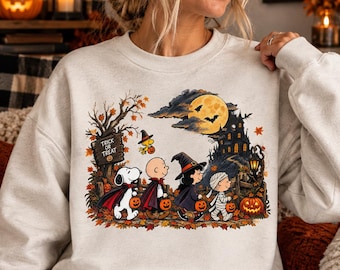 Può includere: Una felpa beige a girocollo con una colorata grafica di Halloween raffigurante personaggi dei cartoni animati in costume, tra cui un vampiro, una strega e una mummia, che camminano davanti a una casa stregata sotto la luna piena con il testo Trick or Treat.