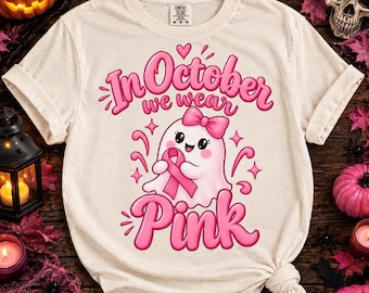 Può includere: Una t-shirt a maniche corte color crema con la grafica di un fantasma bianco con un fiocco rosa, che tiene in mano un nastro di sensibilizzazione rosa. Il testo In October we wear Pink è scritto in grassetto rosa, circondato da stelline rosa.