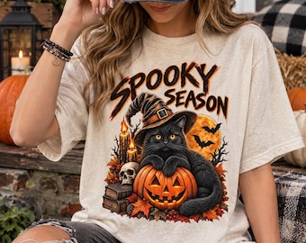 Può includere: Una t-shirt color crema con una stampa grafica che riporta il testo "SPOOKY SEASON" sopra un gatto nero con un cappello da strega, appollaiato su una zucca illuminata. Il design include candele, un teschio, libri, foglie autunnali e pipistrelli.