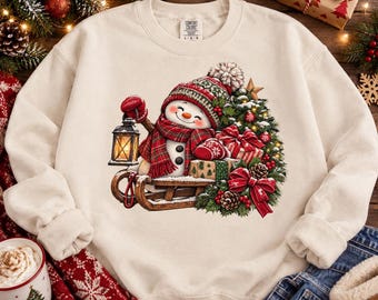 Può includere: Una felpa girocollo color crema con la stampa di un pupazzo di neve sorridente che indossa una sciarpa e un cappello a quadri rossi e verdi. Il pupazzo di neve è seduto su una slitta di legno e tiene in mano una lanterna accesa.