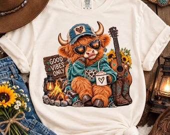 Può includere: Una t-shirt color crema con la grafica di una soffice mucca highland che indossa un berretto blu, occhiali da sole a forma di cuore e una felpa con cappuccio color ottanio. La mucca tiene una tazza con un cuore, accanto a una chitarra e girasoli.