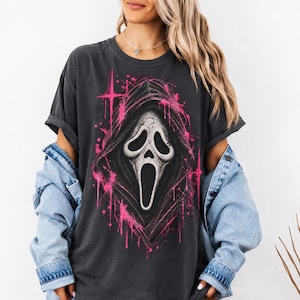 May include: A charcoal gray short-sleeve t-shirt featuring a large graphic print of a white ghost mask with a black hood, surrounded by bright pink star-shaped accents and paint splatter effects, worn with a light blue denim jacket over the shoulders.