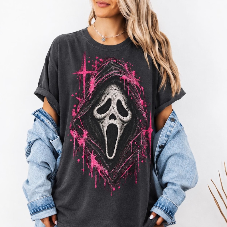 May include: A charcoal gray short-sleeve t-shirt featuring a large graphic print of a white ghost mask with a black hood, surrounded by bright pink star-shaped accents and paint splatter effects, worn with a light blue denim jacket over the shoulders.