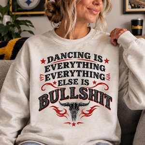 Könnte beinhalten: Ein beigefarbenes Sweatshirt mit Rundhalsausschnitt und schwarzem sowie rotem Text, der DANCING IS EVERYTHING EVERYTHING EVERYTHING ELSE IS BULLSHIT lautet. Das Design enthält rote Sterne, dekorative rote Schnörkel und einen schwarzen Stierschädel.