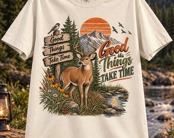Può includere: Una t-shirt a maniche corte color crema con una stampa grafica raffigurante un cervo in una foresta vicino a un ruscello. Il design include un cartello in legno con il testo "Good Things Take Time", una catena montuosa, un sole e uccelli.