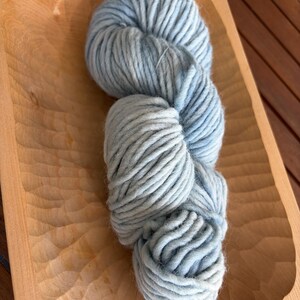 Könnte beinhalten: Ein einzelner Strang aus dickem, grobem Garn in einem sanften, hellblauen Farbton mit subtilen weißen Variationen. Das Garn ist zu einer langen, abgerundeten Form gedreht und weist über die gesamte Länge eine flauschige, weiche Textur auf.
