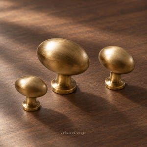 May include: Three oval-shaped cabinet knobs in a brushed brass finish with a metallic sheen. Each knob features a smooth, rounded top and a cylindrical base. The text "VelaireDesign" is printed in small, dark letters at the bottom center.