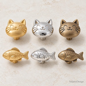 以下が含まれることがあります： 猫の形をしたノブ3つと魚の形をしたノブ3つからなる、金、銀、ブロンズ仕上げの金属製キャビネットノブのセット。画像の右下隅には「VelaireDesign」というテキストが印刷されています。
