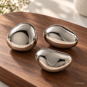 May include: Three smooth, polished silver-toned decorative objects with irregular, rounded pebble-like shapes. Each piece features a highly reflective, mirror-like surface that captures light and surrounding reflections, creating a sleek appearance.