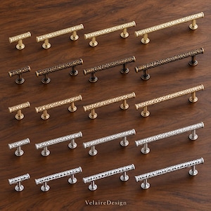 May include: A collection of twenty metal cabinet pulls arranged in five rows, featuring hammered textures in gold, dark bronze, light gold, brushed nickel, and polished chrome finishes. Each pull has two cylindrical posts and the text VelaireDesign.
