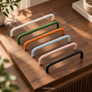 May include: A set of six U-shaped cabinet pulls in white, green, orange, light blue, pink, and black, arranged diagonally on a wooden surface. The text "VelaireDesign" is printed in small, centered letters at the bottom of the wooden board.